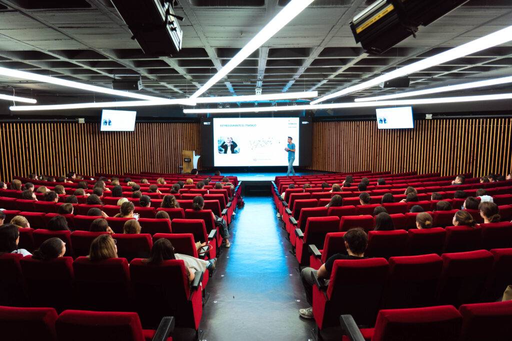 La imagen muestra un auditorio lleno de personas sentadas, con un presentador en el escenario frente a una pantalla.