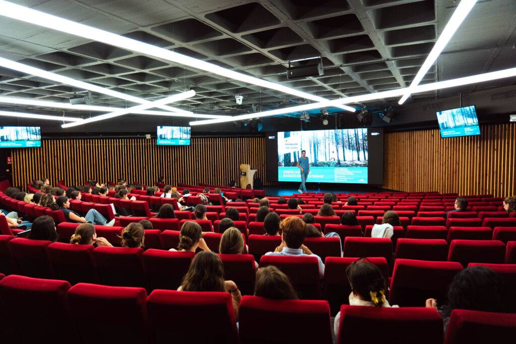 Una sala de conferencias con asientos rojos y un presentador en el escenario frente a una audiencia.