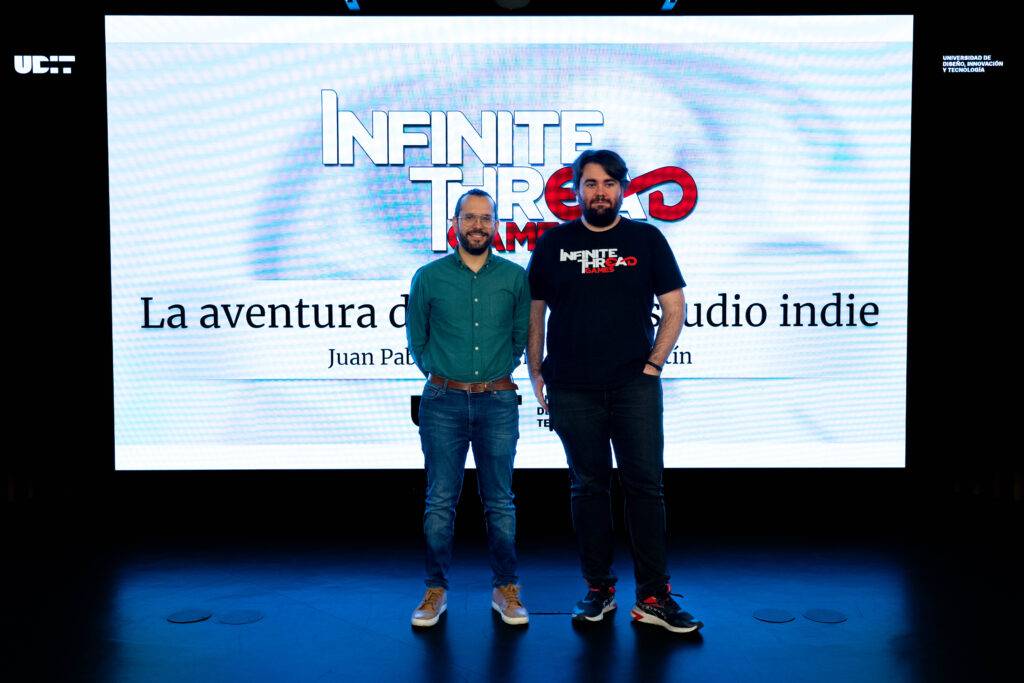 Dos personas posan frente a una pantalla con la presentación de un evento sobre desarrollo de videojuegos indie.