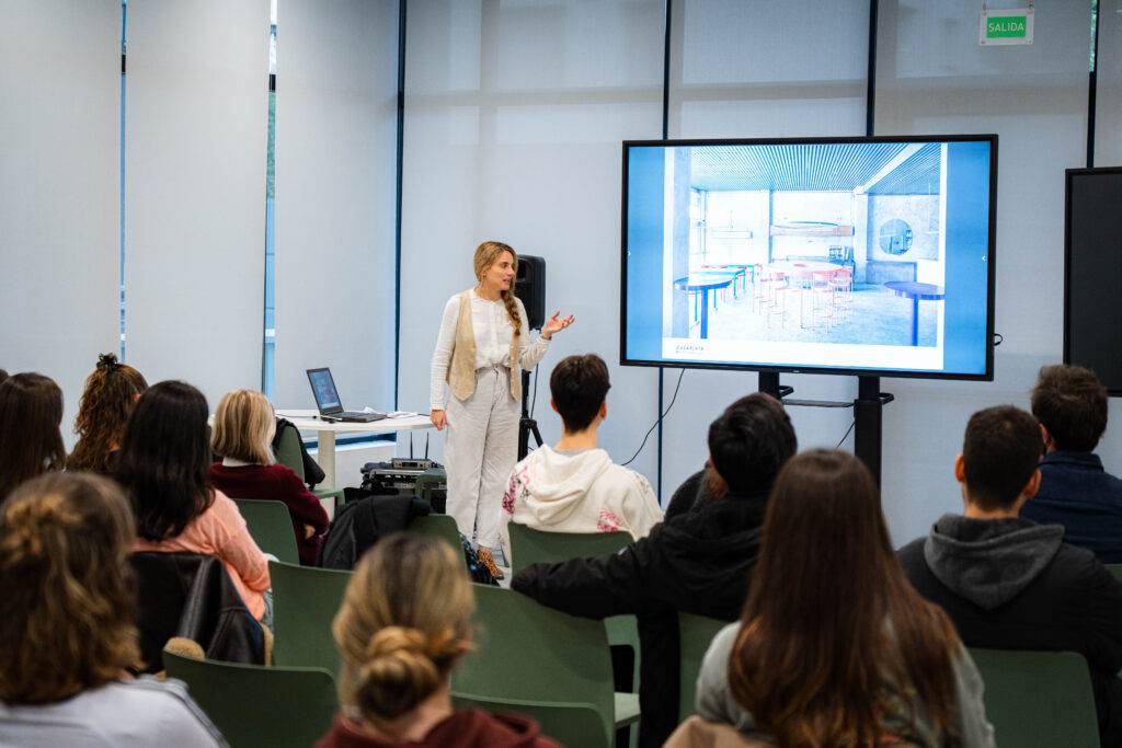 Una presentadora expone ante un público en un evento con una pantalla que muestra una imagen de un ambiente interior.