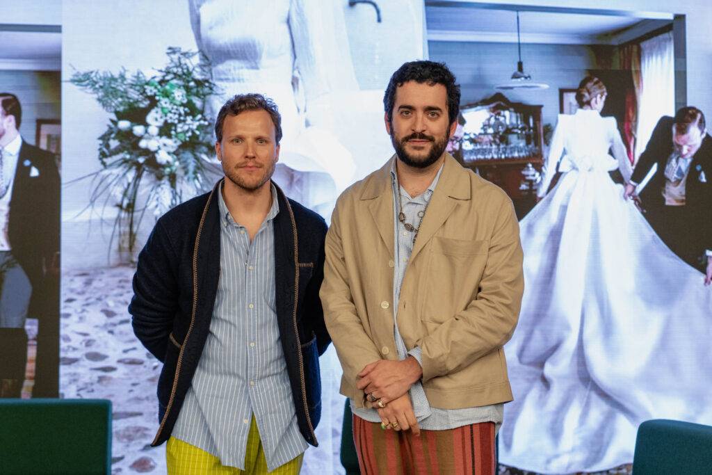 Dos hombres posando frente a una pantalla con una imagen de fondo que muestra una elegante escena de boda.