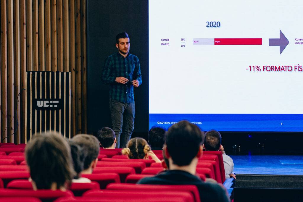 Un presentador habla frente a una pantalla que muestra datos de mercado en una conferencia.