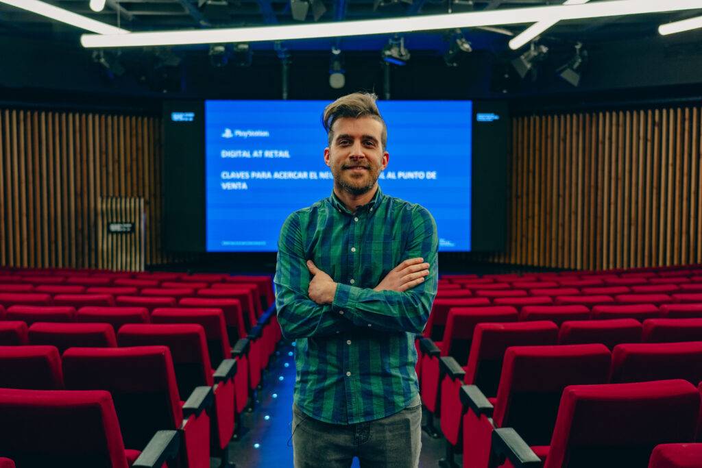 Un hombre sonriente se encuentra de pie frente a un auditorio con asientos rojos y una pantalla grande detrás de él.