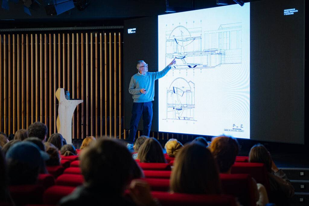 Un ponente presenta un diseño arquitectónico en una sala llena de personas que lo observan.