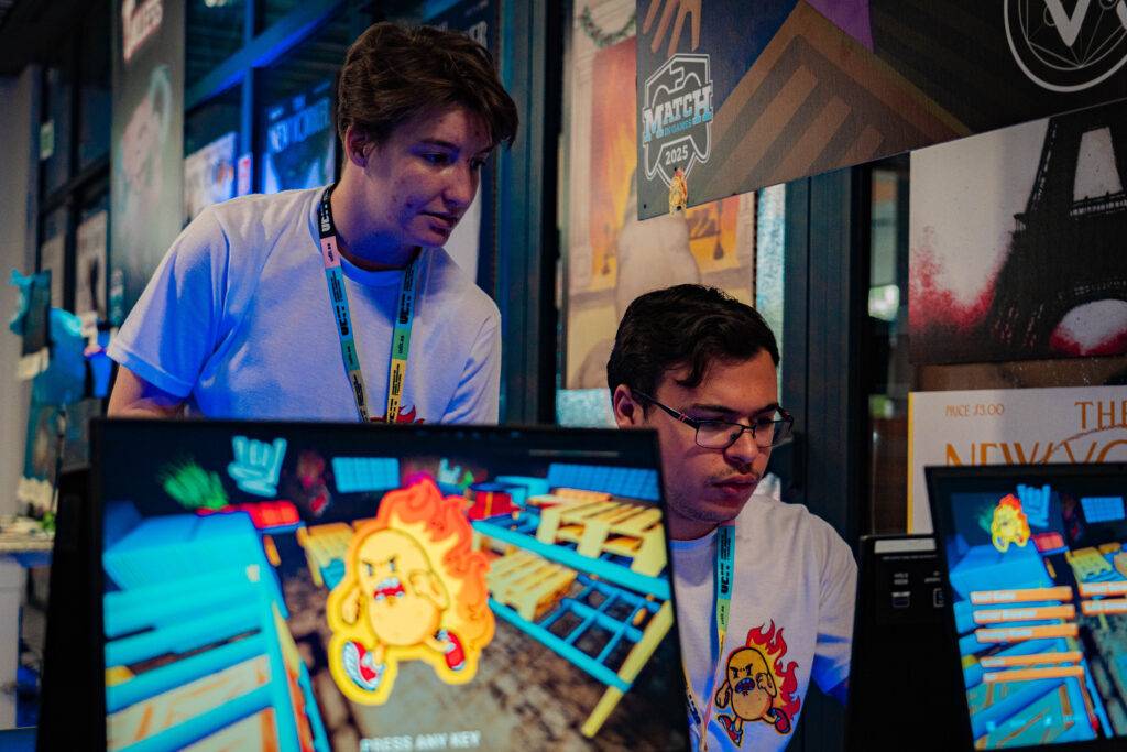Dos jóvenes interactúan en un evento de videojuegos mientras observan una pantalla.