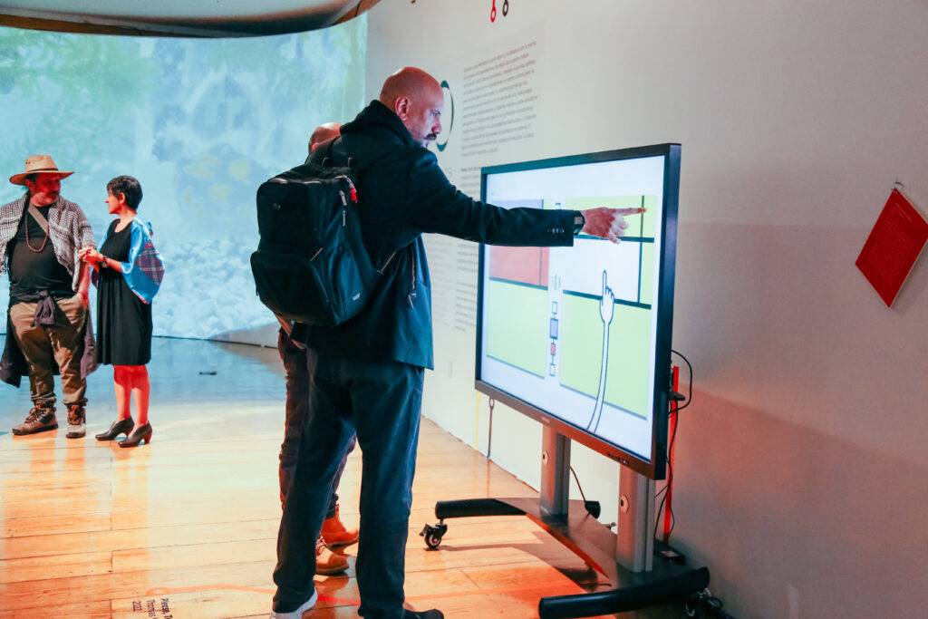 Un hombre interactúa con una pantalla táctil en una galería de arte mientras otros visitantes observan.