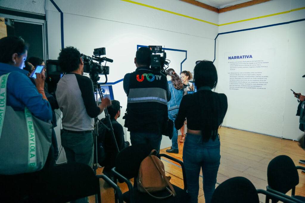 Un grupo de personas con cámaras se reúne en un evento mientras una mujer habla frente a una pared con un texto.