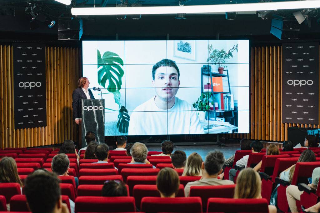 Una presentación en un auditorio donde un ponente habla ante una gran pantalla que muestra a una persona.