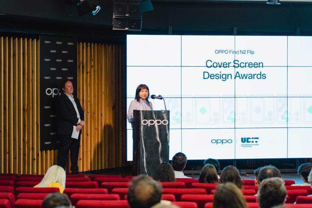 Una mujer habla en un podio durante una ceremonia de premios de diseño en un auditorio con asientos rojos.