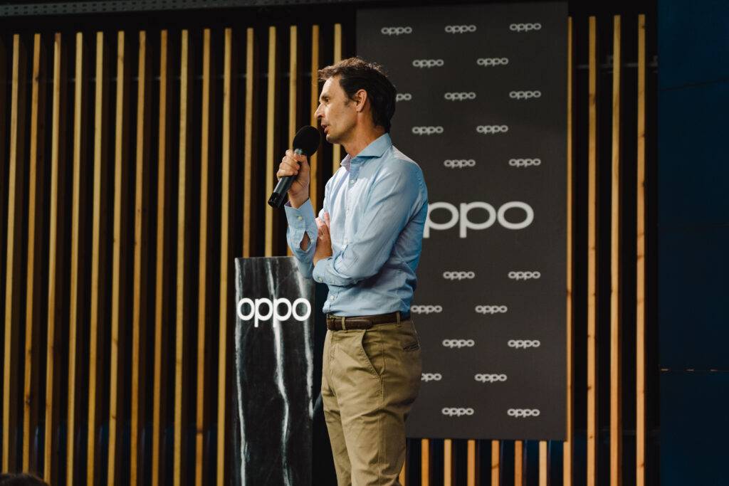 Un hombre está hablando en un evento frente a un panel con el logo de la marca Oppo.