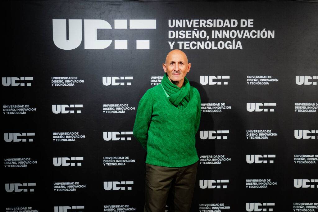 Un hombre con un suéter verde posando frente a un fondo que muestra el logotipo de la Universidad de Diseño, Innovación y Tecnología.