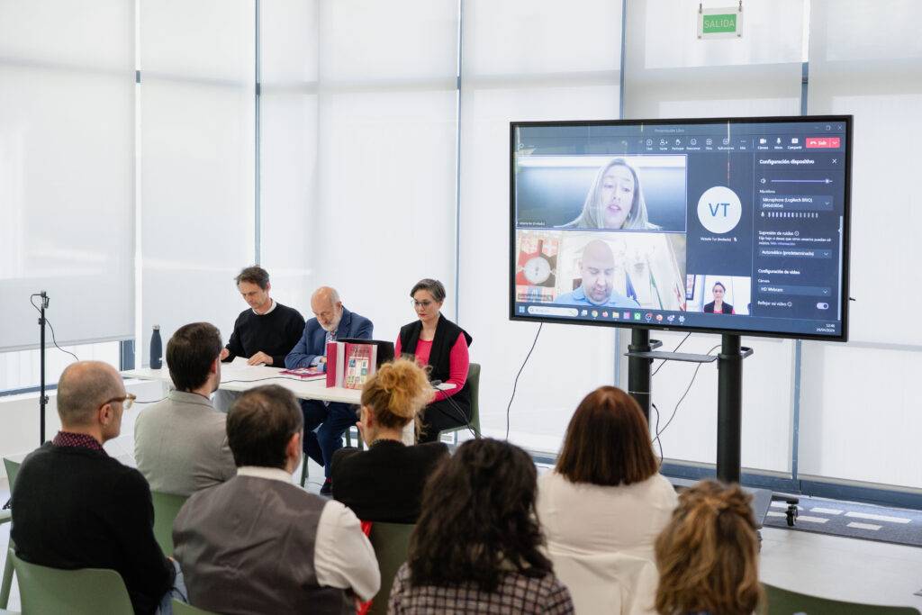 Una mesa de conferencias con varias personas sentadas y una pantalla mostrando una videollamada.