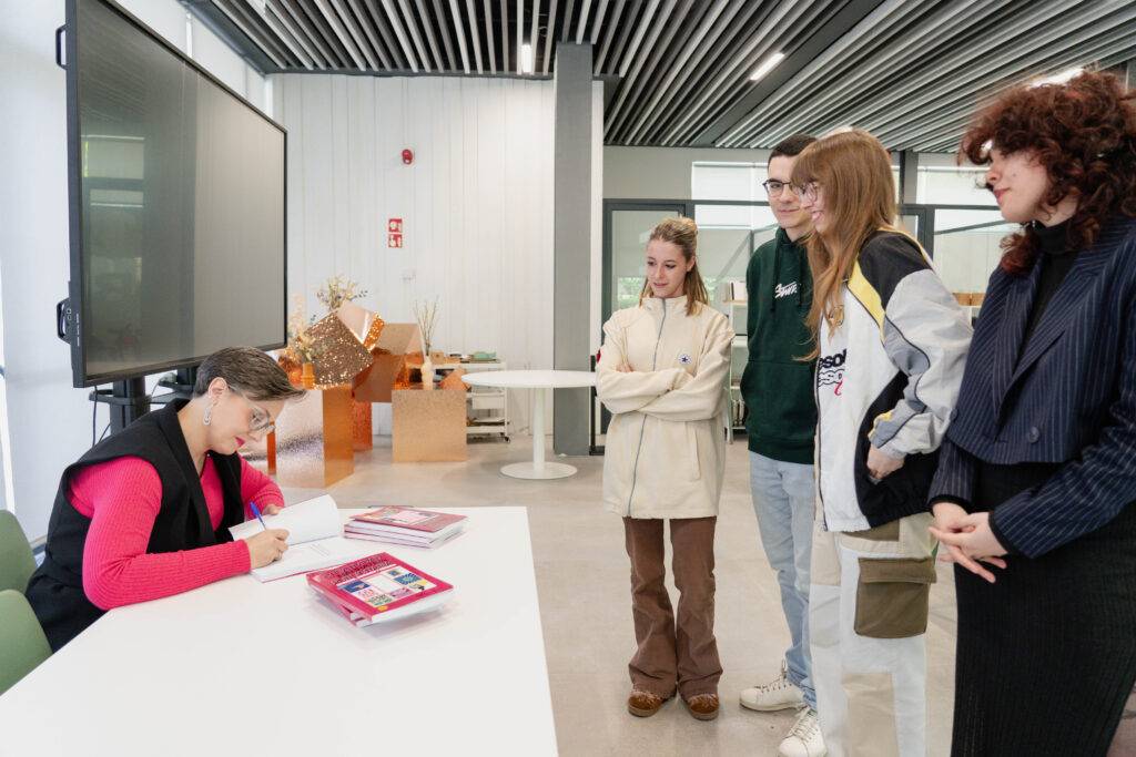 Una autora firma libros mientras un grupo de jóvenes la observa en una biblioteca moderna.