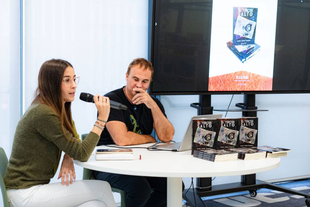 Una presentación de libro en la que una joven habla mientras un hombre escucha atentamente.