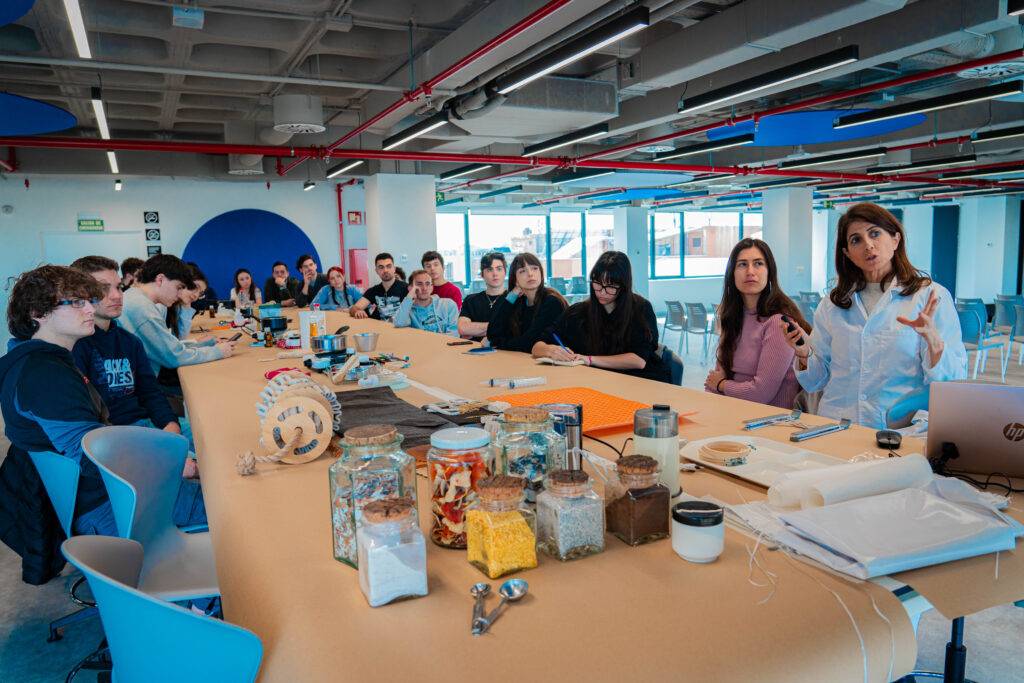 Un grupo de personas se encuentra en una sala de trabajo, escuchando a una mujer que explica algo frente a un largo escritorio lleno de materiales.