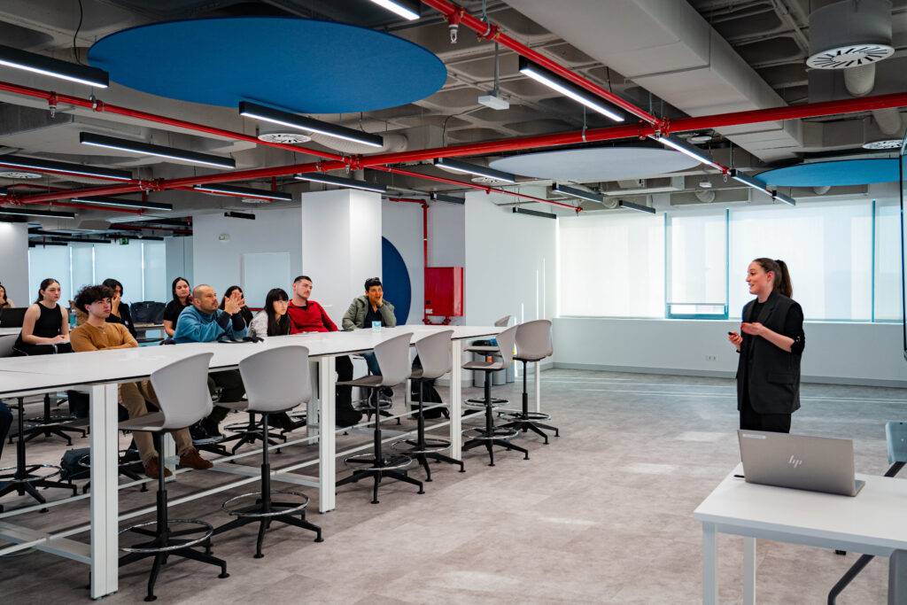 Una presentadora está dando una charla frente a un grupo de personas en un espacio moderno y bien iluminado.