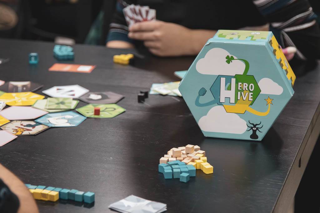 La imagen muestra un juego de mesa sobre una mesa con varias piezas de color y un dado hexagonal con el título 'Hero Hive'.
