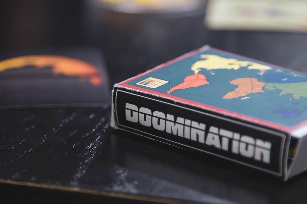 La imagen muestra una caja de un juego de mesa titulado 'DOOMINATION' con un diseño colorido de mapas en su portada.