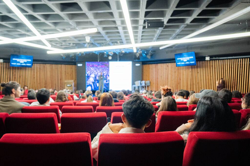 Una sala llena de personas sentadas en asientos rojos, mirando hacia la pantalla en un evento.