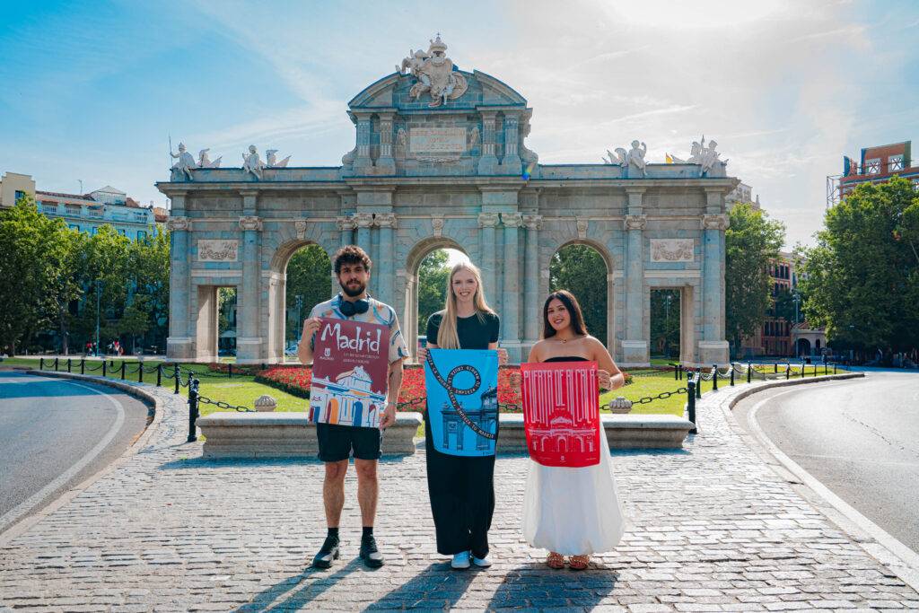 Tres personas posan frente a un monumento en Madrid, cada una sosteniendo una pintura.
