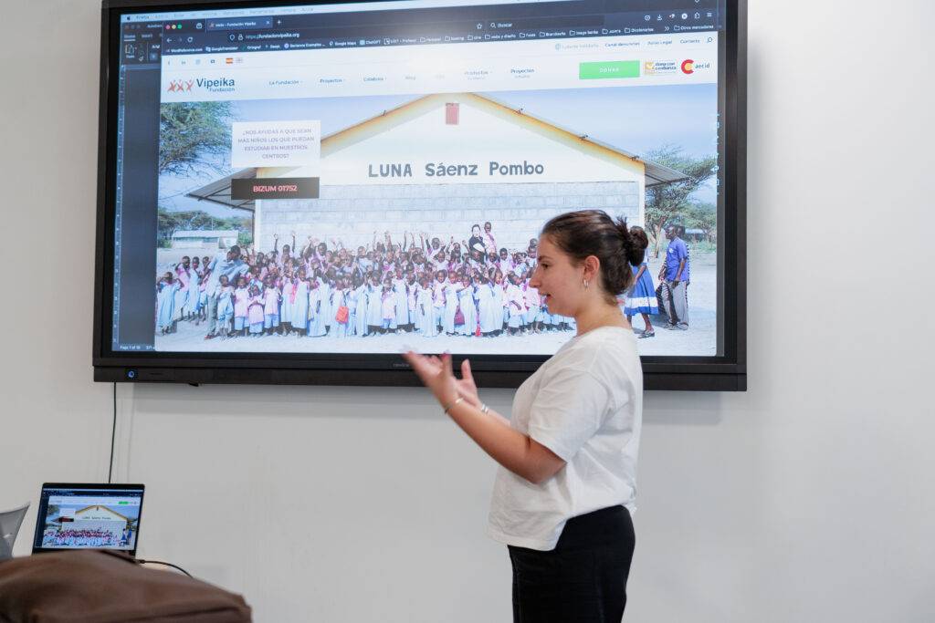 Una persona está presentando información sobre un proyecto educativo frente a una pantalla que muestra a un grupo de niños.