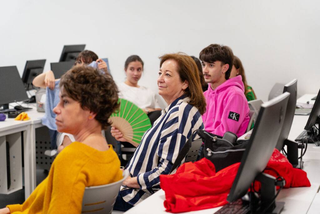 Un grupo de personas sentadas en un aula, algunas mirando hacia la cámara mientras una mujer sostiene un abanico.
