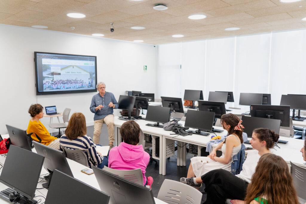 Una clase en un aula con un profesor explicando frente a un grupo de estudiantes y computadoras.
