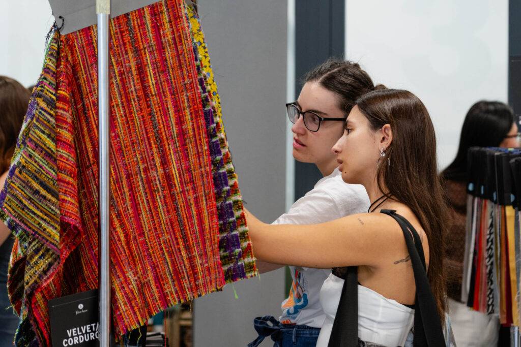 Dos mujeres observan y tocan piezas de tela en un evento.
