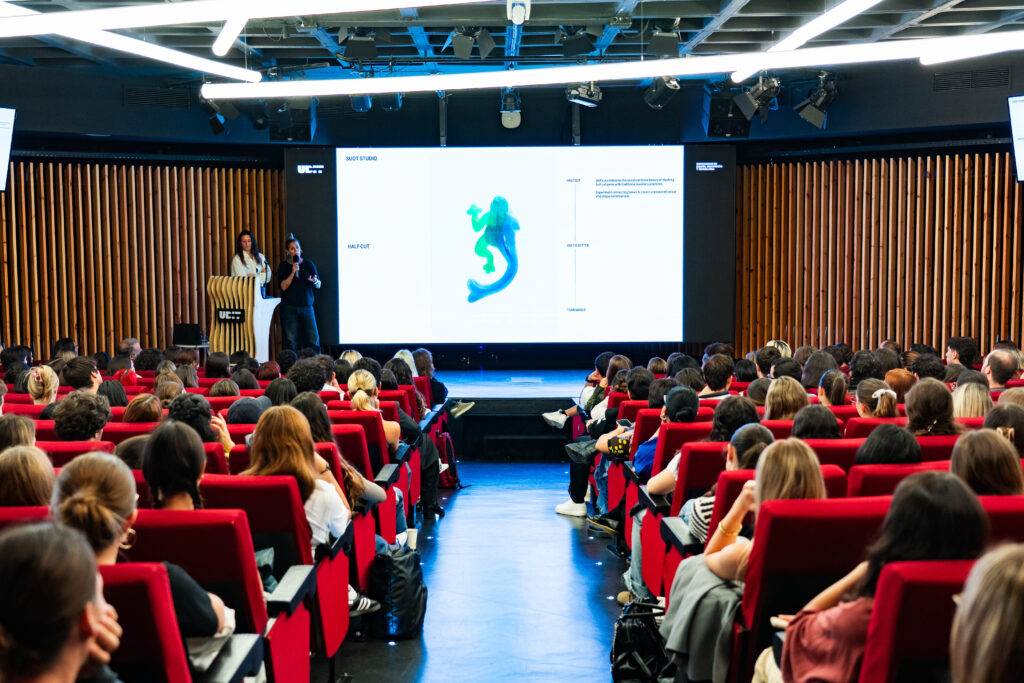 Una conferencia con un gran público sentado en un auditorio y dos ponentes en el escenario.