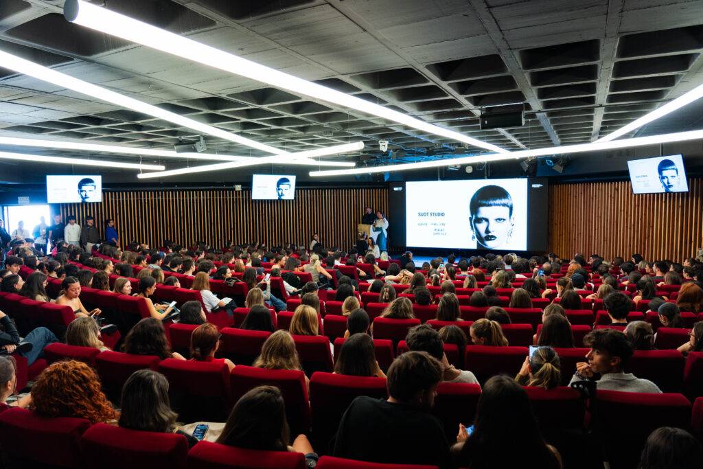 Una sala de conferencias llena de gente sentada, con pantallas mostrando una presentación en el fondo.