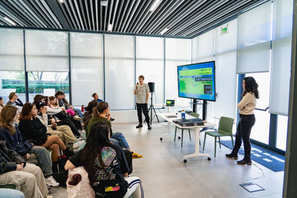 Una charla educativa en un aula moderna donde dos presentadores muestran información a un público atento.