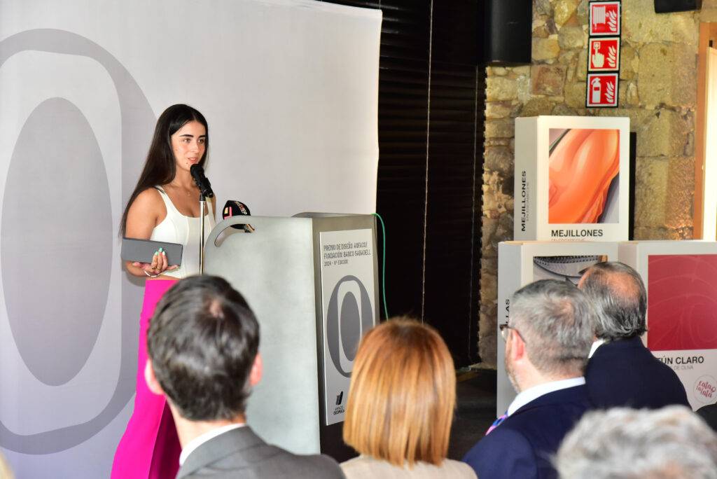 Una mujer joven habla en un evento frente a un podio con un micrófono, mientras un grupo de personas la escucha.