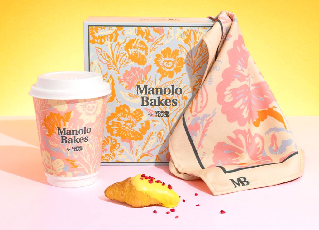 Un conjunto de productos de repostería de la marca Manolo Bakes, que incluye una taza de café, una caja decorada y un croissant.