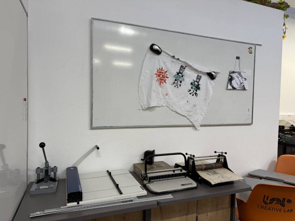 Una mesa de trabajo con herramientas de impresión y una camiseta colgada en una pizarra blanca.