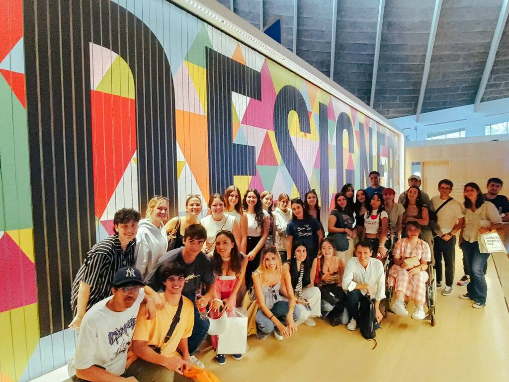 Un grupo de personas posando frente a un mural colorido con la palabra 'DESIGN' en un espacio interior.