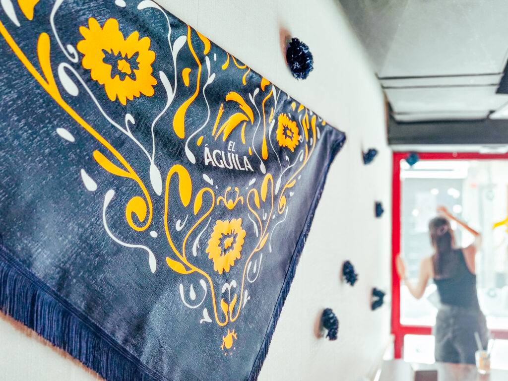 Una bandana decorativa con flores amarillas y el texto 'El Águila' en un ambiente luminoso.