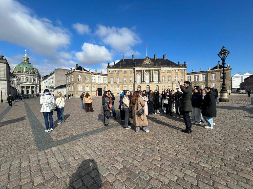 Un grupo de personas se encuentra en una plaza frente a un edificio histórico bajo un cielo azul con algunas nubes.