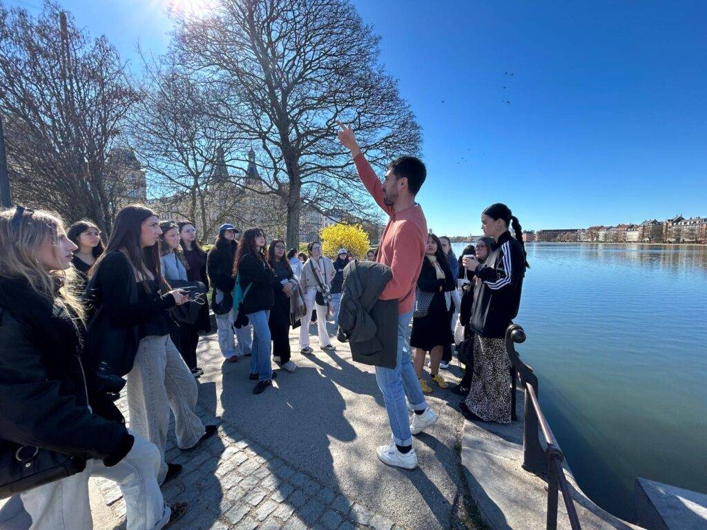 Un grupo de personas escucha a un hablante junto a un río en un día soleado.