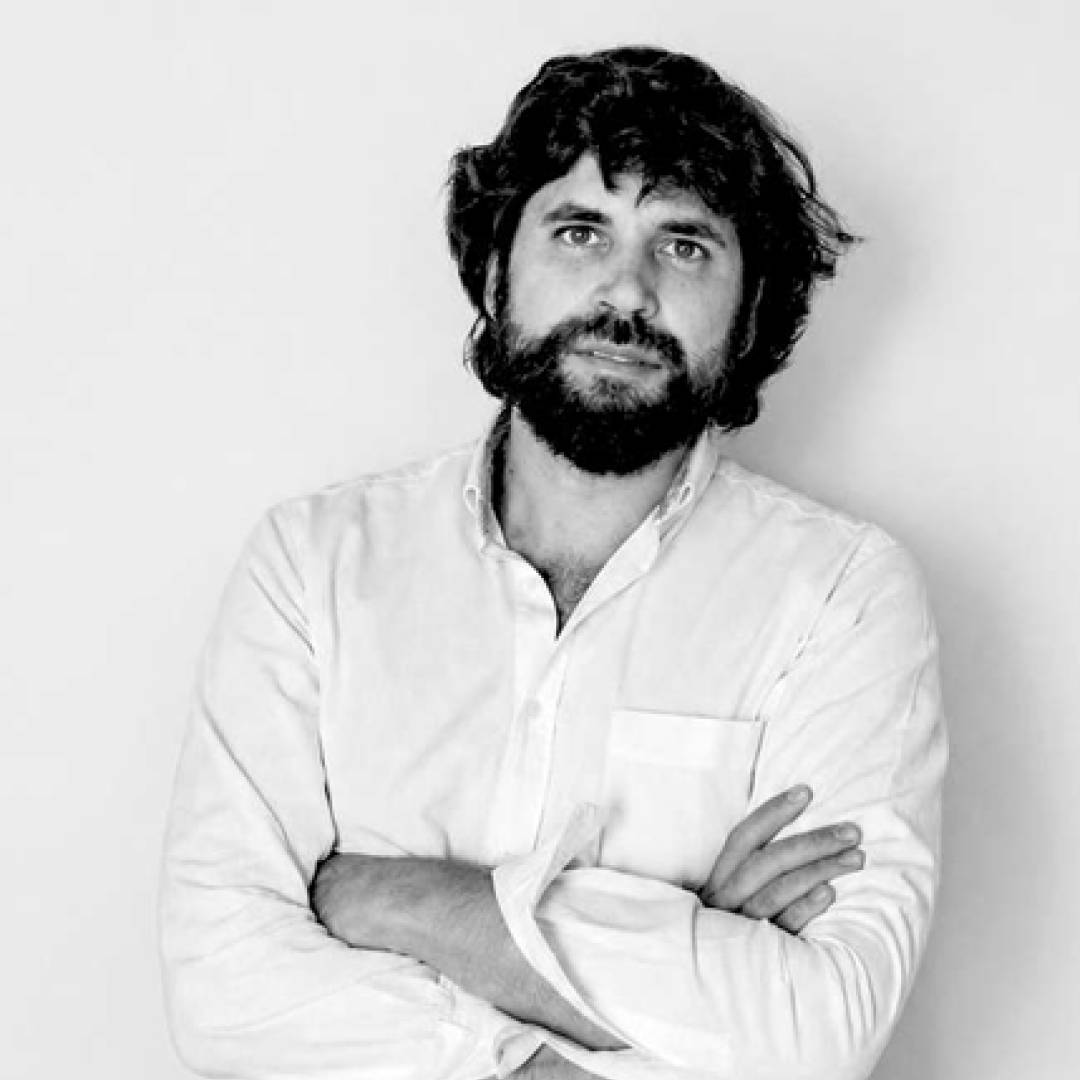 Un hombre con barba y cabello rizado viste una camisa blanca y se apoya en una pared blanca con los brazos cruzados.