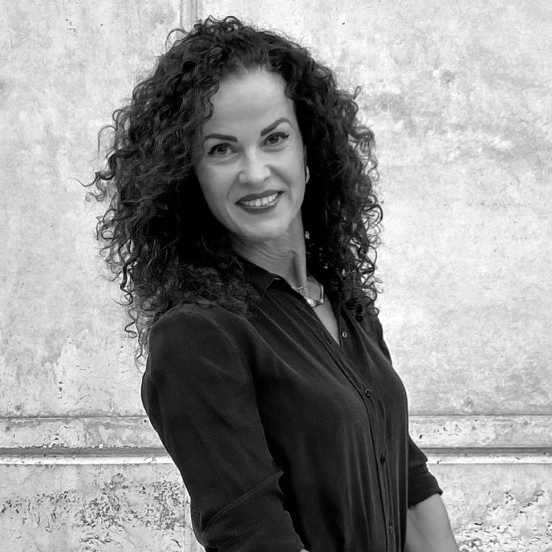 Una mujer sonriente con cabello rizado y una blusa negra frente a un fondo claro.