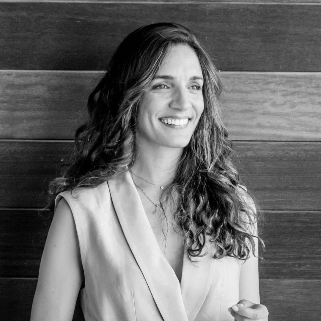 Una mujer sonriente con cabello rizado se presenta frente a un fondo de madera oscura.