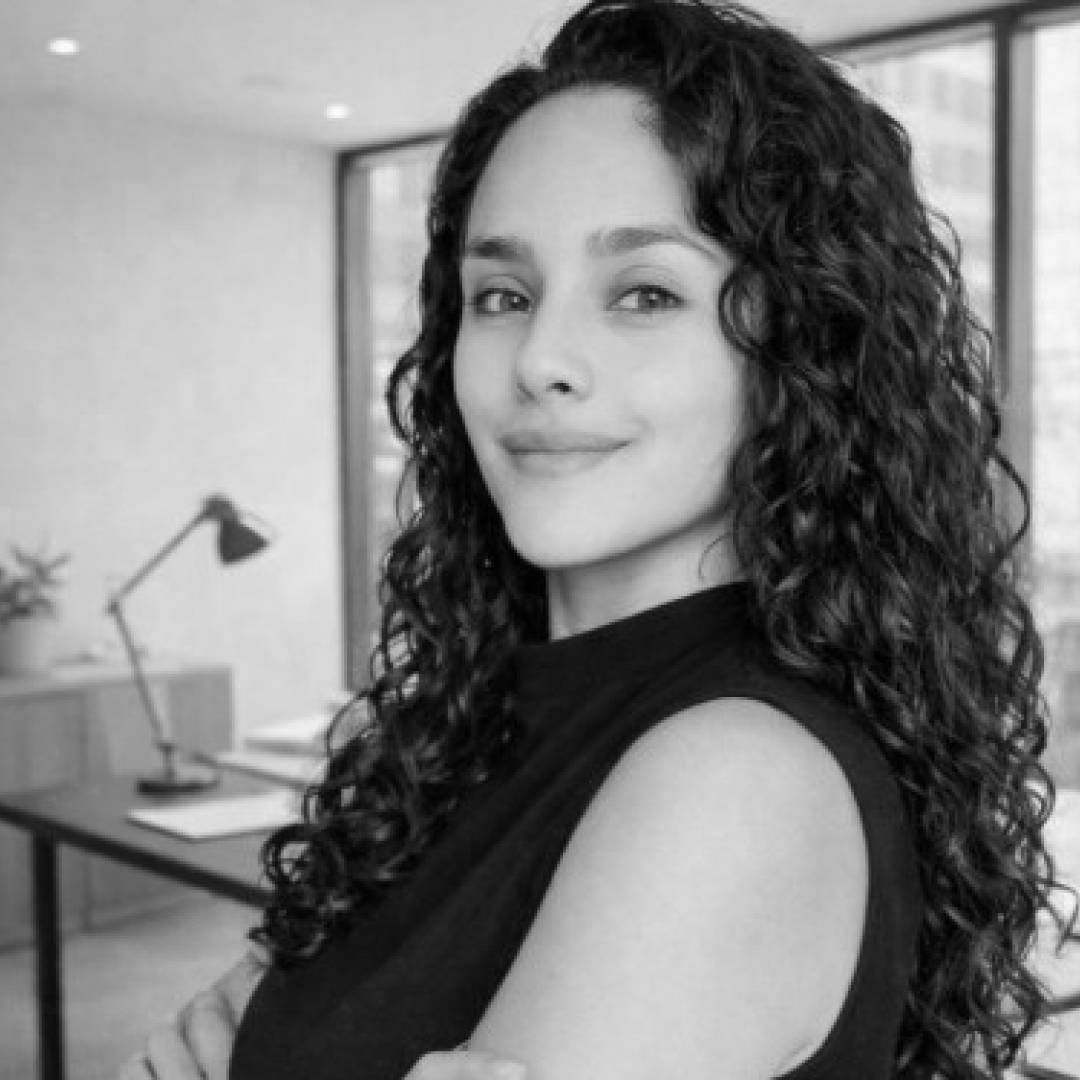 Una mujer sonriente con cabello rizado se encuentra en una oficina moderna.