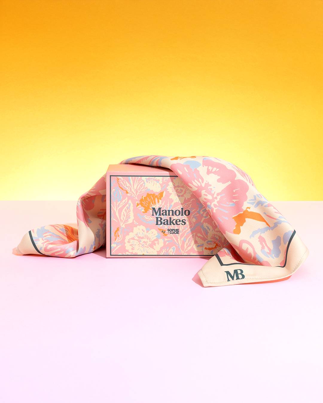 Una caja floridamente decorada de Manolo Bakes sobre una superficie rosa, acompañada de una bufanda elegante.