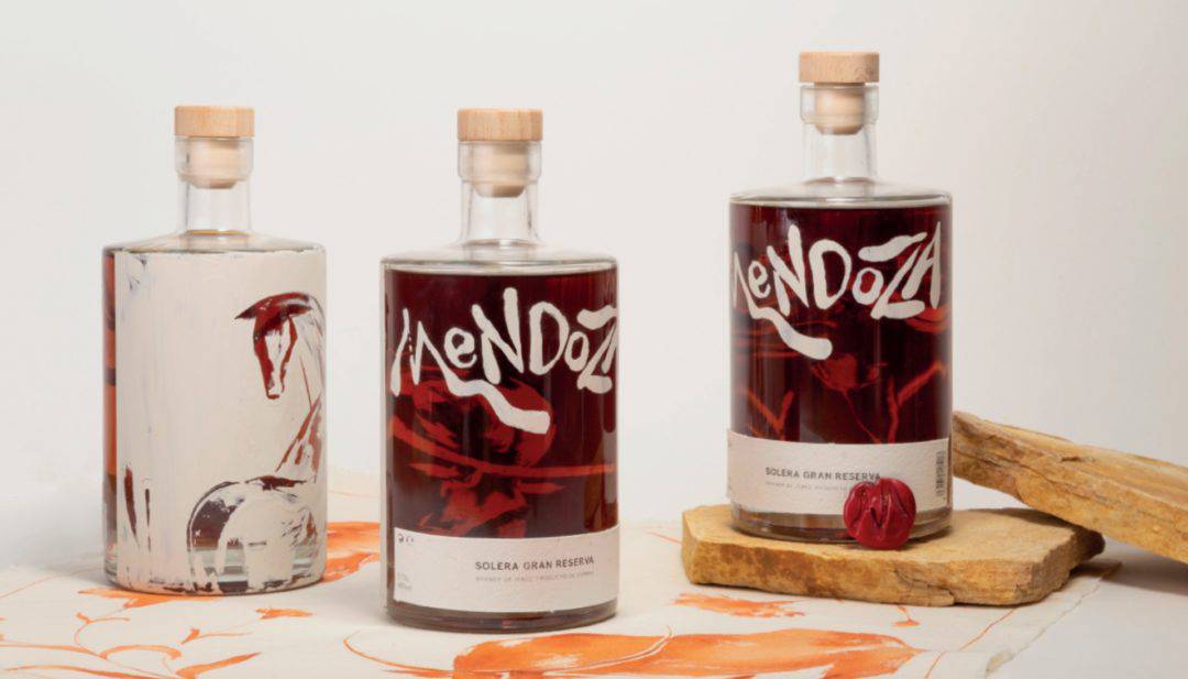 Tres botellas con etiquetas artísticas de una bebida alcohólica sobre una mesa de madera clara.