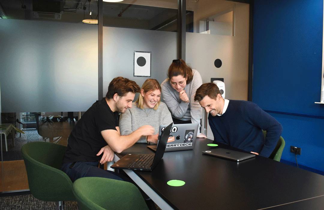 Un grupo de cuatro personas se reúne alrededor de una laptop en un ambiente de oficina, mostrando interés en lo que están viendo en la pantalla.