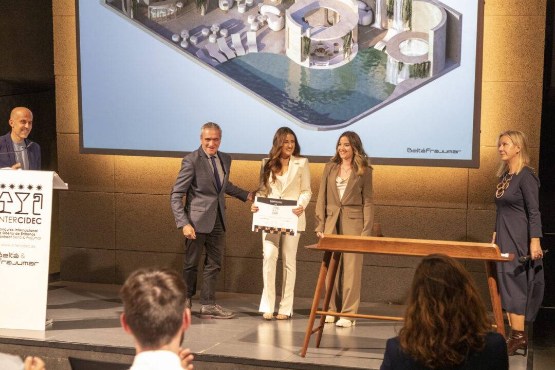 Un grupo de personas está en un evento, entregando un premio en un escenario con un fondo arquitectónico.