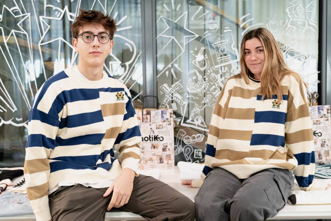 Dos jóvenes posan juntos luciendo suéteres de rayas en un ambiente moderno.