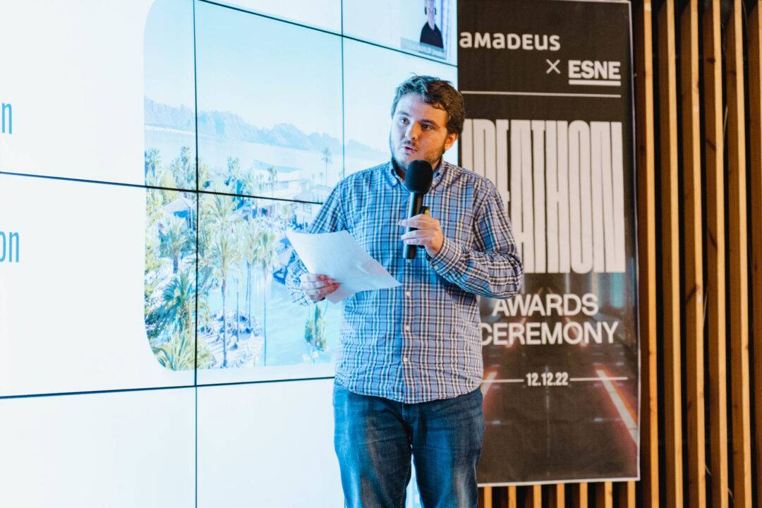 Un hombre presenta en un evento frente a una pantalla con imágenes y un cartel de ceremonia de premios.