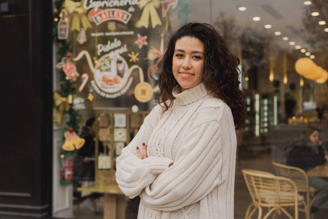 Una joven sonriente con un suéter blanco posando enfrente de una cafetería decorada para la temporada.
