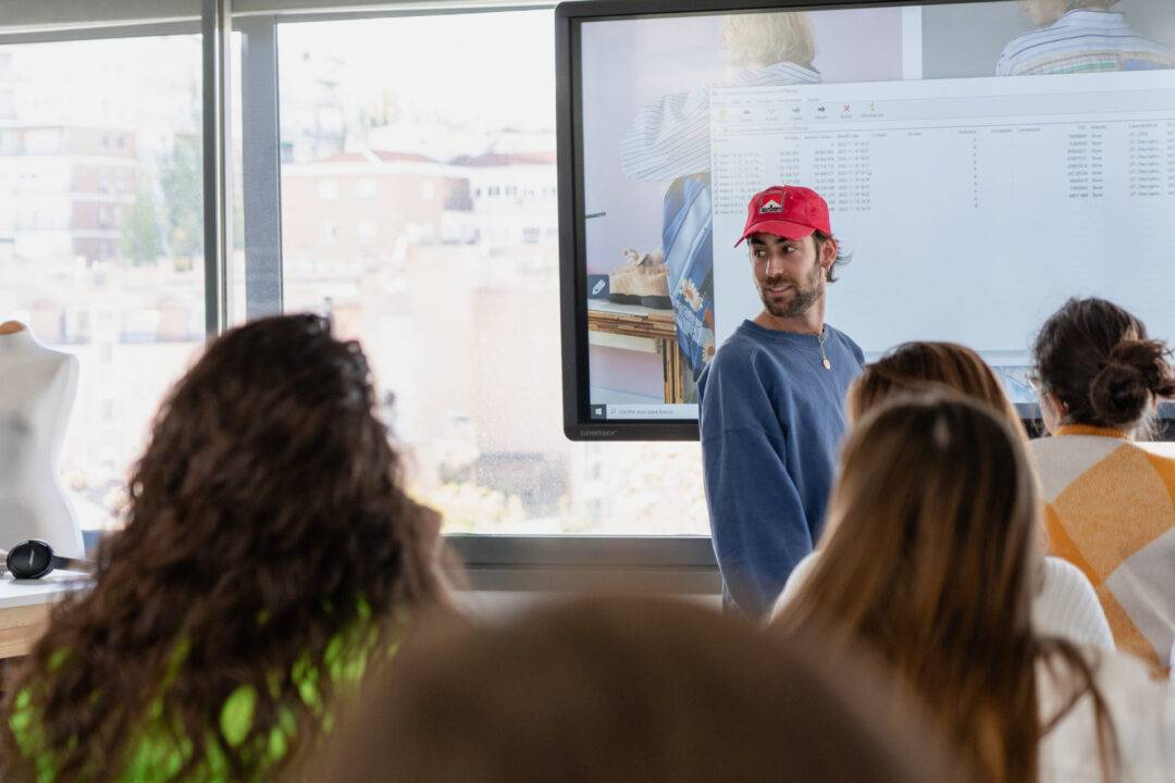 Un hombre con gorra roja explica algo frente a un grupo de personas en un aula.